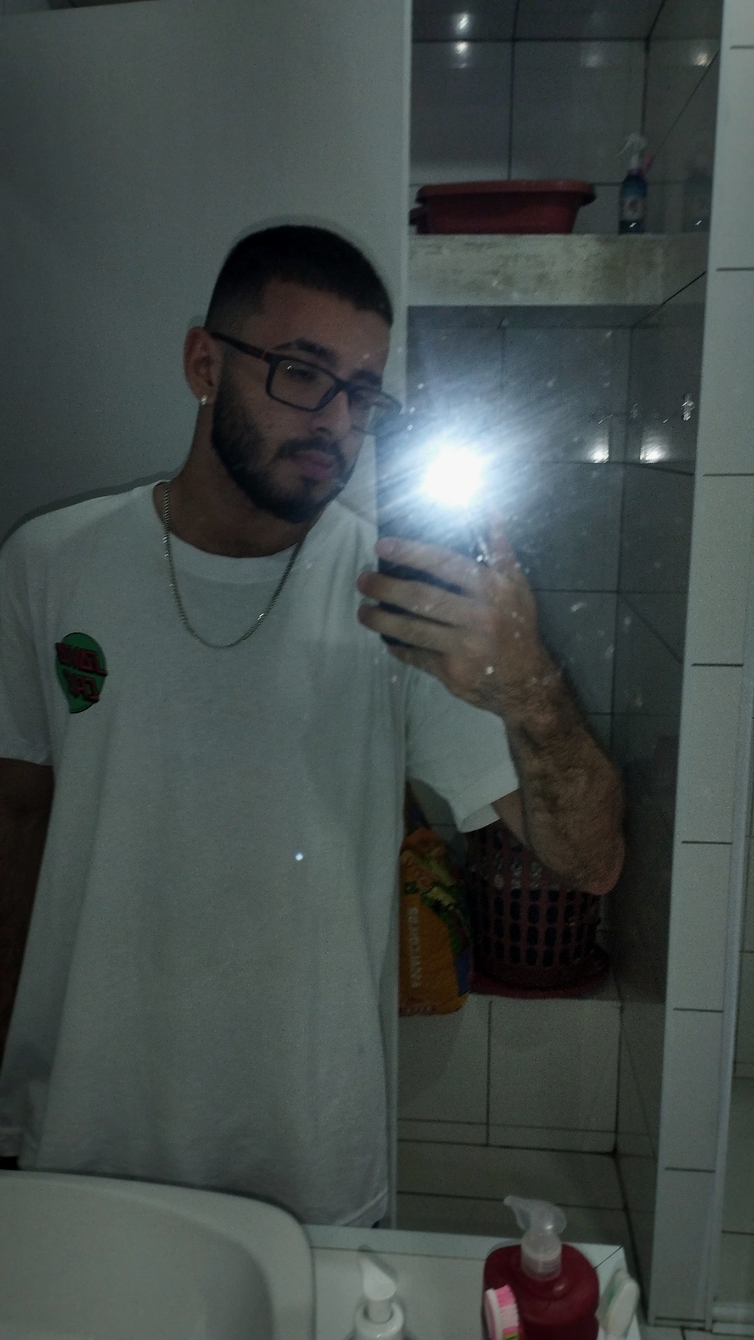 Selfie (com flash) de Igor no banheiro. Ele está de camiseta branca, cabelo curto (máquina), barba aparada, camiseta branca e corrente